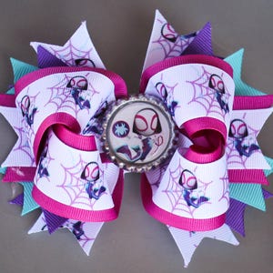 May include: A handmade hair bow with a Spider-Gwen theme. The bow is made with white ribbon printed with spiderwebs and the character, accented with pink and purple ribbons. A Spider-Gwen bottle cap centre adds detail. The bow is a fun accessory.