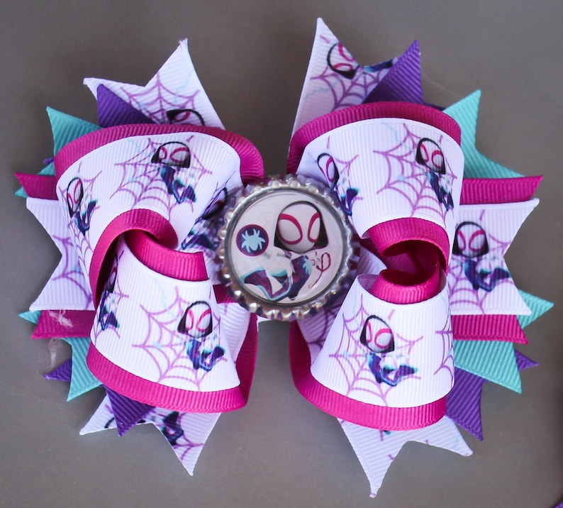 May include: A handmade hair bow with a Spider-Gwen theme. The bow is made with white ribbon printed with spiderwebs and the character, accented with pink and purple ribbons. A Spider-Gwen bottle cap centre adds detail. The bow is a fun accessory.