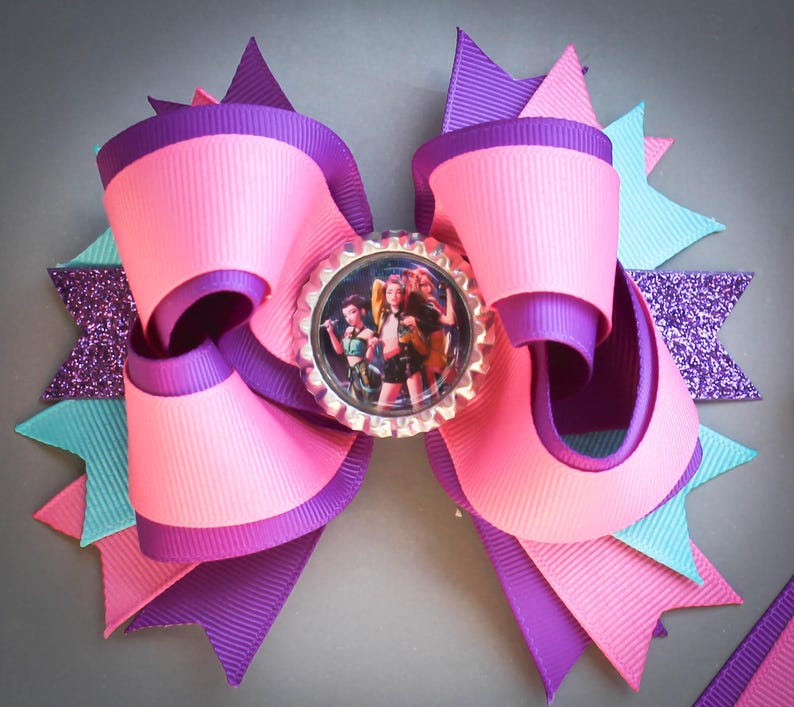 May include: A large, multi-layered hair bow in shades of pink, purple, and turquoise. The bow features a central image within a bottle cap, surrounded by ribbon loops and glitter accents. The bow is designed with a layered effect, creating a visually appealing accessory.