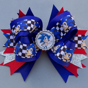 K&ouml;nnte beinhalten: Handgefertigte Haarschleife im Sonic the Hedgehog-Design. Die Schleife ist &uuml;berwiegend blau, rot und wei&szlig;, mit einem Schachbrettmuster und einem zentralen Sonic-Bild. Akzente sind silberner Glitzer und ein Flaschenverschluss als Mittelst&uuml;ck.