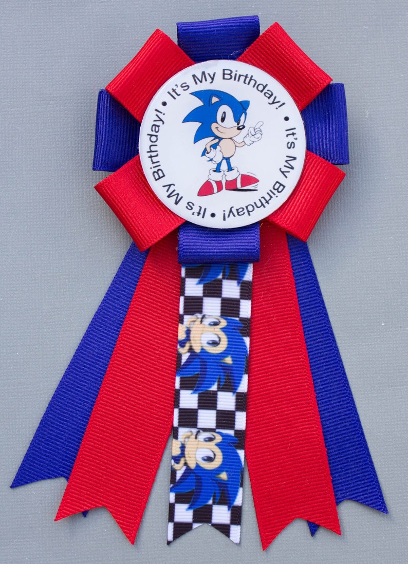 K&ouml;nnte beinhalten: Eine festliche Geburtstagsrosette mit dem Charakter Sonic the Hedgehog. Die Rosette hat rote und blaue B&auml;nder, mit einem zentralen runden Emblem mit der Aufschrift "It's My Birthday!". Ein kariertes Band mit Sonics Bild ist ebenfalls vorhanden.