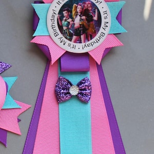 May include: A colorful birthday ribbon with a central image of three people. The ribbon features pink, purple, and teal accents, a glittery bow, and the text "It's My Birthday!" around the image.