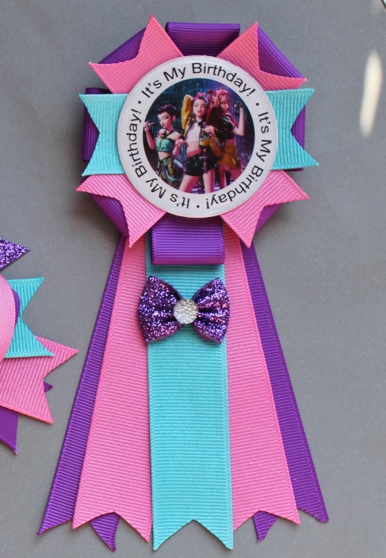 May include: A colorful birthday ribbon with a central image of three people. The ribbon features pink, purple, and teal accents, a glittery bow, and the text "It's My Birthday!" around the image.