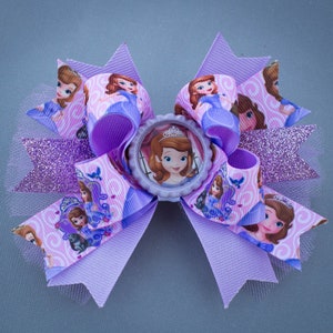 May include: Purple and pink hair bow with a princess character in a purple dress and crown. The bow has a glittery purple center and a white tulle accent.
