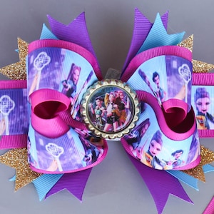 May include: A large, layered hair bow in shades of pink, purple, and blue, with gold glitter accents. The bow features a central image of a group of people, printed ribbon, and a bottle cap centerpiece, creating a decorative accessory.