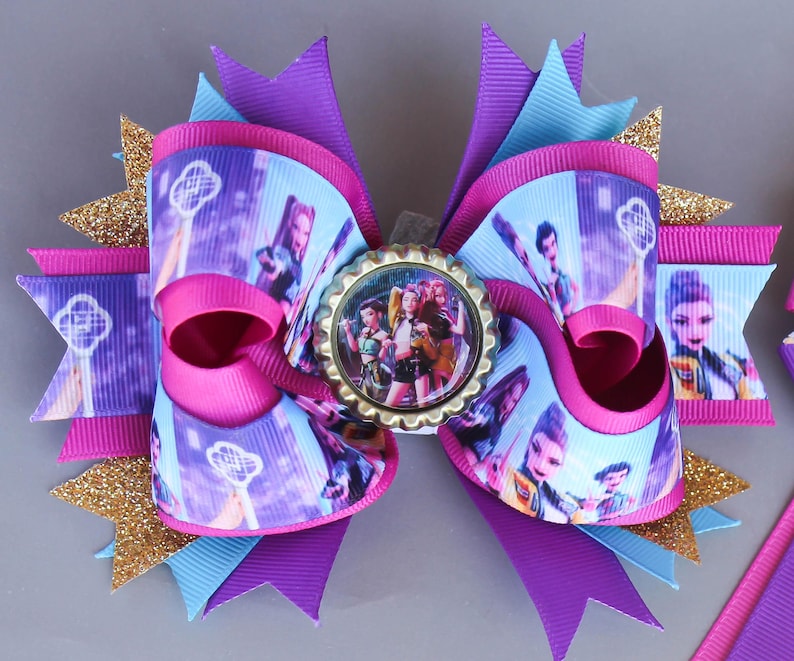 May include: A large, layered hair bow in shades of pink, purple, and blue, with gold glitter accents. The bow features a central image of a group of people, printed ribbon, and a bottle cap centerpiece, creating a decorative accessory.