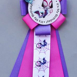May include: A birthday ribbon with a Spider-Gwen theme. The ribbon is predominantly pink and purple, with a white ribbon displaying a web pattern and Spider-Gwen graphics. The centre has a circular design with the text "It's My Birthday!" and a glittery bow.