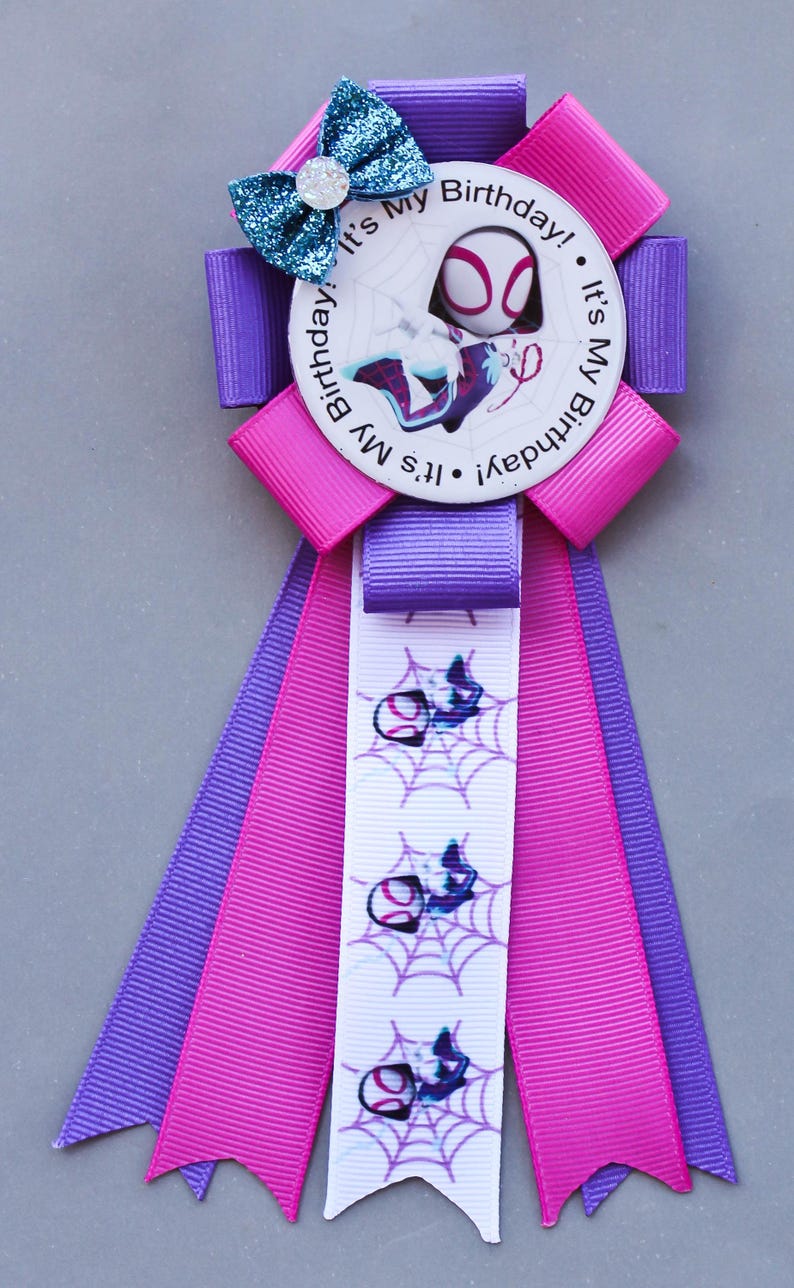 May include: A birthday ribbon with a Spider-Gwen theme. The ribbon is predominantly pink and purple, with a white ribbon displaying a web pattern and Spider-Gwen graphics. The centre has a circular design with the text "It's My Birthday!" and a glittery bow.