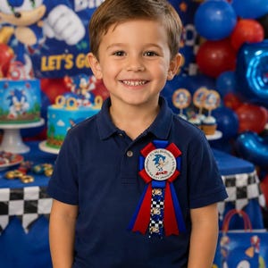 K&ouml;nnte beinhalten: Ein l&auml;chelnder Junge in einem marineblauen Poloshirt und khakifarbener Hose. Er tr&auml;gt eine Geburtstagsrosette mit Sonic the Hedgehog. Der Hintergrund zeigt Sonic-Dekorationen, Luftballons und eine Geburtstagstorte, was eine festliche Atmosph&auml;re schafft.