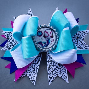 May include: A turquoise and white bow with a spiderweb design. The center of the bow features a silver bottle cap with a cartoon image of Spider-Gwen.