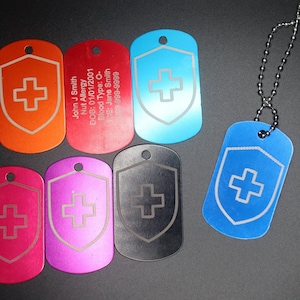 May include: Six different coloured metal dog tags with a white cross inside a shield design. One tag has the text "John J Smith", "Nut Allergy", "DOB: 01/01/2001", "Blood Type: O-", "CG: Jane Smith", "100-999-9999". A silver chain is attached to one of the tags.
