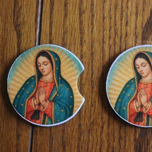 May include: Two round car coasters featuring an image of Our Lady of Guadalupe. The image shows a woman with hands clasped in prayer, wearing a blue cloak and red robe, surrounded by a golden halo. The coasters have a white border.
