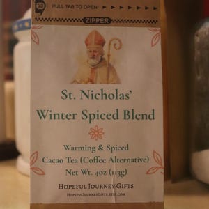 May include: A brown paper bag of St. Nicholas' Winter Spiced Blend cacao tea. The bag features an illustration of St. Nicholas and text that reads "Warming & Spiced" and "Net Wt. 4oz (113g)".