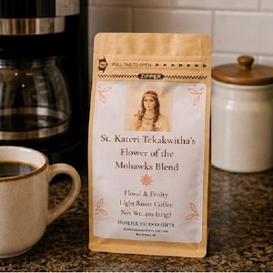Könnte beinhalten: Eine Packung hell gerösteten Kaffees "St. Kateri Tekakwitha's Flower of the Mohawks Blend" mit floralem und fruchtigem Geschmacksprofil. Die Packung befindet sich neben einer Kaffeetasse und einer Kaffeemaschine. Das Nettogewicht beträgt 113 Gramm.