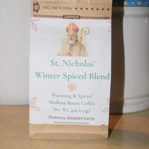 May include: A brown paper bag of St. Nicholas' Winter Spiced Blend medium roast coffee. The bag features an illustration of St. Nicholas and text that reads "Warming & Spiced". Net weight is 4 oz.