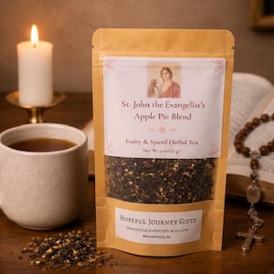 Könnte beinhalten: Eine Packung Kräutertee von St. John the Evangelist's Apple Pie Blend. Der braune Papiersack hat ein klares Fenster, das die Teemischung zeigt. Eine brennende Kerze, eine Tasse Tee und ein Rosenkranz sind ebenfalls im Bild.