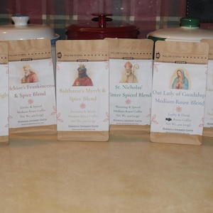 May include: Six coffee bags with various blends, including "Caspar's Golden Light Roast Blend" and "St. Nicholas Inter Spiced Blend." Each bag features a different religious figure illustration and text. The bags are brown with white labels and floral accents.