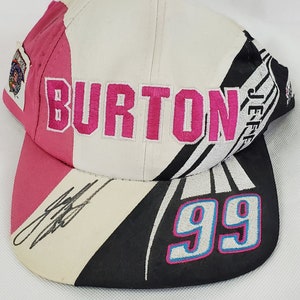 May include: A white, pink, and black baseball cap with the word "BURTON" embroidered in pink on the front. The number "99" is embroidered in white with pink and blue outlines on the back of the cap. The cap is signed in black marker.