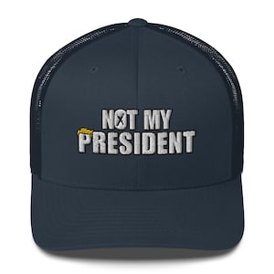 May include: A navy blue trucker hat with a white embroidered message that reads "NOT MY PRESIDENT" with a yellow X over the word "MY".