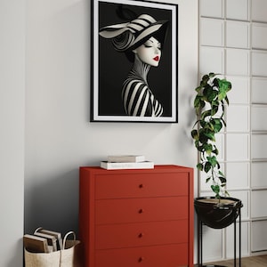 May include: A framed wall art print featuring a stylized portrait of a person wearing a black and white striped hat and matching high-neck garment, accented with bright red lips, displayed above a solid red four-drawer chest of drawers.