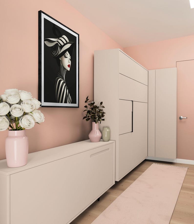 May include: A framed wall art print featuring a stylized portrait of a person in a black and white striped hat and outfit with red lips, displayed in a room with a light pink cabinet, a pink vase with white roses, and a tall beige storage unit.