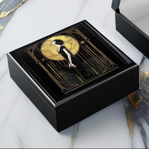 May include: Black wooden jewellery box with a glossy finish. The lid features a gold and black Art Deco design of a woman in a long dress against a golden sunburst background. The box is rectangular and has a hinged lid.