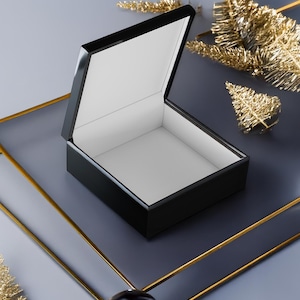 May include: An open, black lacquered box with a white interior sits on a gray surface with gold trim. The box is rectangular with a hinged lid. Gold decorative elements are scattered around the box.