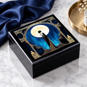 May include: A square black wooden box featuring a decorative lid with a graphic illustration of a figure in a long gown standing before a large yellow moon against a deep blue background, framed by intricate gold-colored geometric line patterns.
