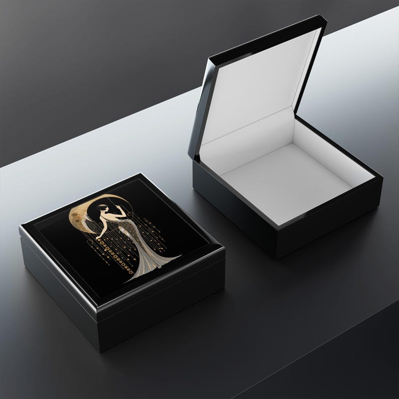 May include: Black rectangular jewelry box with a glossy finish. The lid features a gold and white art deco design of a woman with a crescent moon. The interior is white. The box is open, revealing the inside.
