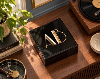 May include: A square, black, high-gloss wooden box featuring the gold-colored, stylized letters AB on the lid. The box has a smooth, reflective finish and a clean, geometric design, showcasing a modern aesthetic with sharp, metallic gold lettering.