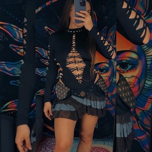 May include: A black long-sleeved top with cut-out detailing and a high neckline. The top has a lace-up design on the front and cut-out sleeves. It is paired with a layered skirt in shades of brown and blue, and a patterned belt.