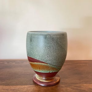 May include: A ceramic goblet with a matte sage green upper section and a glossy deep red base. The middle portion features a textured, horizontal band of burnt orange and brown glaze that separates the two primary colours of the vessel.