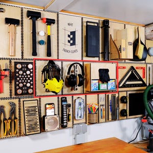 May include: A wall-mounted storage system with various tools and items organized on wooden shelves. The shelves are labeled "Neat French Cleats". The system features a variety of tools, including hammers, a pair of headphones, a small radio, and a set of rulers.