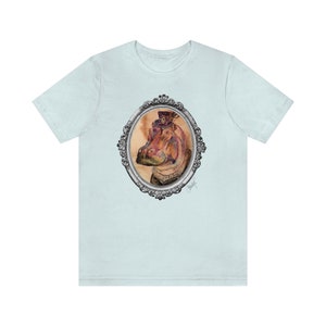 Puede incluir: Una camiseta azul claro con una ilustración de acuarela de un hipopótamo con un sombrero de copa y un collar decorativo. El hipopótamo está enmarcado en un marco ovalado plateado con detalles ornamentados. La firma del artista es visible en la esquina inferior derecha del marco.