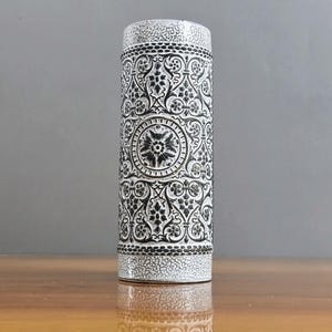 May include: A cylindrical ceramic vase featuring a white and black colour scheme. The surface is decorated with intricate black floral and scrollwork patterns, contrasted by textured white bands at the top and bottom edges of the vessel.