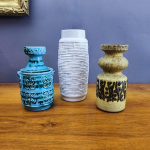 May include: Three ceramic vases of varying shapes and textures. The left vase is bright blue with dark blue embossed scroll patterns. The centre vase is tall, white, and features a raised basket-weave texture. The right vase is tan with dark brown speckles.