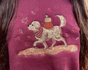 May include: A maroon sweatshirt with an embroidered design of a golden retriever dog carrying a stack of pumpkins. The dog is wearing a red and white plaid scarf. The design is surrounded by small embroidered leaves and stars.