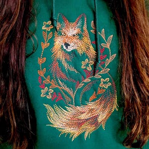 May include: A dark green hoodie with a detailed embroidered fox design in shades of orange, yellow, and red. The fox is surrounded by floral elements.