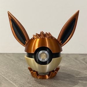 May include: A 3D printed figurine of a Pokemon character, Eevee, in a Poké Ball. The figurine is made of copper-colored plastic and has black ears and a white Poké Ball.