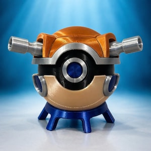 May include: A 3D-printed, multi-colored object resembling a futuristic device. It features a metallic orange top, silver accents, a black center with a blue circle, and two silver cylindrical protrusions. The base is blue and has a tripod design.