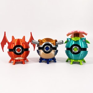 May include: Three colorful, spherical 3D-printed figures. The figures are red, gold, and teal, each with a black band and a central circular design. The red figure has wing-like extensions, the gold one has side tubes, and the teal one has a flower.
