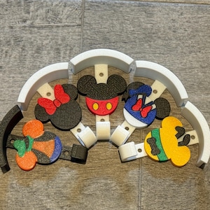 May include: A set of colorful, 3D-printed wall hooks featuring Mickey Mouse, Minnie Mouse, and other Disney character designs. The hooks are attached to a white, semi-circular base, perfect for a child's room or Disney-themed space.