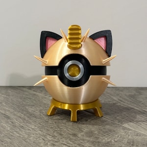 May include: A gold-colored, spherical decorative object with black accents, cat ears, and spikes. The object has a black band around the middle and sits on a gold-colored, three-legged stand. The ears are black with pink interiors.