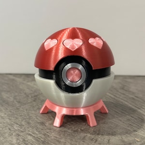 May include: A red and white Poke Ball with pink hearts on the top and a pink base. The Poke Ball is a popular collectible toy.
