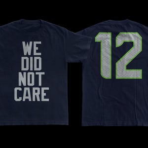May include: Navy blue t-shirt with the text "WE DID NOT CARE" on the front. The back of the shirt displays the number "12" in a silver and green design. The shirt is made of a soft material.