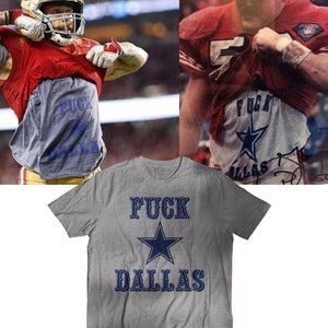 May include: Gray t-shirt with the text "FUCK DALLAS" in navy blue, with a navy blue star graphic. The image also shows two football players wearing jerseys and revealing the same text on their undershirts.