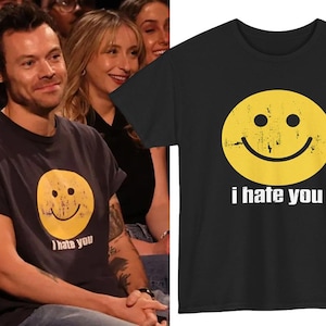 May include: Black t-shirt with a distressed yellow smiley face and the white text "i hate you". The t-shirt is a crew neck style. The image shows the t-shirt being worn by a person. The material appears to be soft.