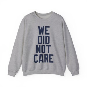 May include: A heather gray sweatshirt with the navy blue text "WE DID NOT CARE" vertically stacked. The long-sleeved crew neck top is made of a soft material and is suitable for casual wear.
