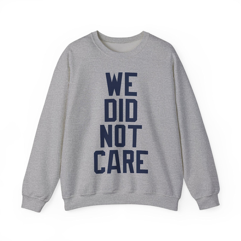 May include: A heather gray sweatshirt with the navy blue text "WE DID NOT CARE" vertically stacked. The long-sleeved crew neck top is made of a soft material and is suitable for casual wear.