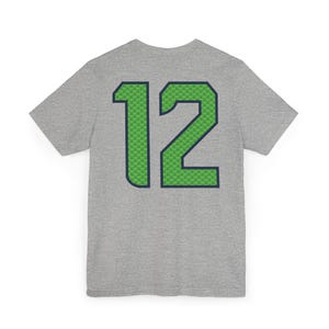May include: A heather gray t-shirt with the number "12" on the back. The number is green with a blue outline and a green pattern fill. The shirt is short-sleeved and has a crew neck.